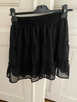 Svart volangkjol med prickig mesh - Snygg svart kortkjol med volanger och transparent prickig mesh längst ner. Resår i midjan för skön passform. Perfekt att styla med en enkel topp eller stickad tröja.