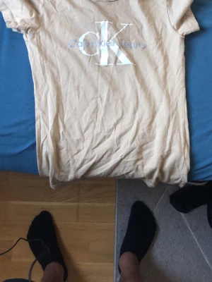 Beige t-shirt från Calvin Klein Jeans - Snygg beige t-shirt från Calvin Klein Jeans med klassisk CK-logga framtill. T-shirten har korta ärmar och är gjord i mjuk bomull som känns skön mot huden. Perfekt till jeans eller shorts för en clean och enkel look.