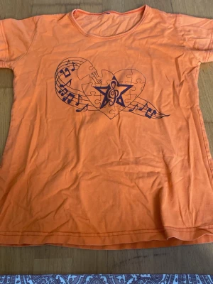 Orange t-shirt med musiktryck - Snygg orange t-shirt med korta ärmar och ett coolt tryck av ett hjärta med noter och stjärna på bröstet. Perfekt för dig som gillar musik och vill sticka ut lite extra. Tillverkad i mjuk bomull för skön känsla hela dagen.