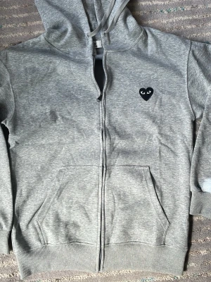Grå hoodie från Comme des Garçons - Säljer en grå hoodie med dragkedja från Comme des Garçons Play. Hoodien har ett svart hjärta med ögon broderat på bröstet, två fickor fram och justerbar huva med snören. Tillverkad i mjuk bomullsblandning, perfekt för en chill och stilren look. Passar bra om du är 175 cm