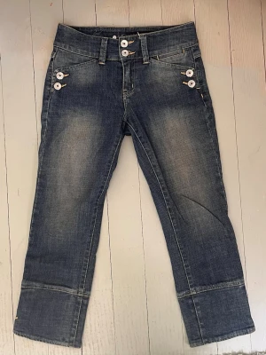 Blå capris från Karen Millen - Säljer ett par blå capris från Karen Millen i super fint skick. Jeansen har dekorativa silver knappar vid fickorna och breda benslut. Klassisk femficksmodell i 100% bomullsdenim med gul kontrastsöm.