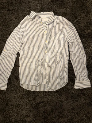 Randig linneskjorta från Zara - Säljer en vit och blå randig skjorta från Zara i skönt linnematerial. Skjortan har klassisk krage, bröstficka och knappar framtill. Perfekt för dig som gillar en avslappnad men ändå stilren look.