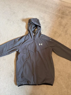 Under Armour löparjacka/vindjacka med huva – Ny med lappar – Strl M - .Säljer en helt ny Under Armour löpar-/vindjacka i storlek M. Jackan är oanvänd och prislapparna sitter kvar. Pris går alltid att diskutera  • Storlek: M • Färg: Mörkgrå • Lätt och smidig vindjacka med huva • Dragkedja framtill och sidofickor med dragkedja • Reflekterande detaljer – perfekt för löpning och träning