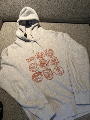 Maison Kitsuné hoodie - Snygg ljusgrå hoodie från Maison Kitsuné med brunt tryck av olika emblem och text på bröstet. Hoodien har huva med dragsko och ribbade muddar vid ärmar och nederkant. Perfekt för en avslappnad och trendig stil.