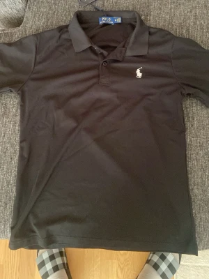Svart skjorta från Polo Ralph Lauren - Klassisk svart pikétröja från Polo Ralph Lauren med vit broderad logga på bröstet. Tröjan har krage, knappar framtill och korta ärmar. Perfekt till en clean och stilren look.