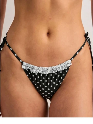 Svart prickig bikiniunderdel med spets - Snygg svart bikiniunderdel med vita prickar och söt vit spetskant framtill. Modellen har knyt i sidorna för justerbar passform och en cheeky baksida. Självklart helt oanvänd, förpackning och allt sitter kvar. Säljer även toppen!