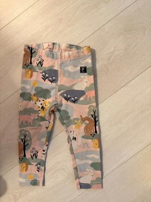 Mönstrade leggings från Polarn O. Pyret - Säljer ett par gulliga leggings från Polarn O. Pyret i storlek 86. De har ett färgglatt djurmönster med kaniner, lejon och träd i pastellfärger. Tillverkade i mjuk ekologisk bomull med justerbar midja och bekväm passform.