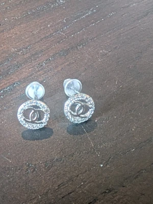 Cc örhängen med strass - Säljer ett par snygga CCl örhängen i silverfärg med rund form och glittrande strass runt logotypen. Klassisk CC-logga i mitten och stift med transparent bakstycke. Perfekta för dig som gillar exklusiva accessoarer.