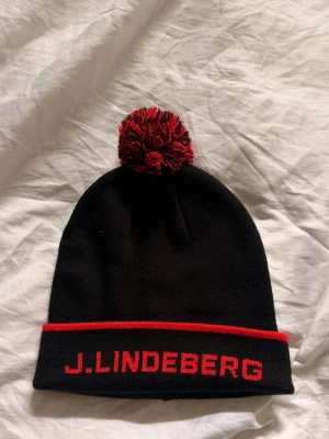 Svart mössa J.Lindeberg - Snygg svart mössa från J.Lindeberg med röd text och en röd rand längs kanten. Perfekt för kalla dagar och ger en sportig look.