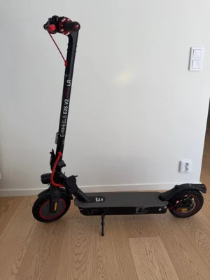Elsparkcykel E-Wheels E25 V2 PRO LR  - Säljer en svart elsparkcykel E-Wheels E25 V2 med röda detaljer. Modellen har bred ståplatta, stora hjul och handtag med röd accent. Perfekt för smidig transport i stan. Laddningsuttag och stöd finns på sidan. 