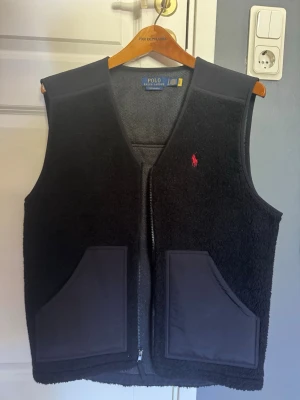 Svart fleeceväst från Polo Ralph Lauren - Svart fleeceväst från Polo Ralph Lauren med dragkedja framtill, två stora fickor och röd broderad logga på bröstet. Västen har en stilren design med mjukt teddy-material och detaljer i slät textil. Perfekt för lager-på-lager och streetwear.