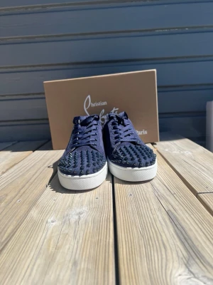 Marinblå sneakers från Louboutin - Snygga marinblå sneakers från Christian Louboutin med klassiska nitar på tån och sidorna. Skorna har en vit sula och är gjorda i mocka med matchande marinblå skosnören. Perfekta för dig som vill sticka ut med en edgy och exklusiv stil.