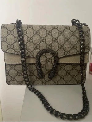 Helt ny! Beige axelväska  - HELT NY! Säljer en handväska i beige GG Supreme canvas med mörkbruna detaljer. Väskan har en kraftig kedjerem i metall och ett dekorativt spänne framtill med ormmotiv. Klassisk rektangulär form och ikoniskt GG-mönster över hela väskan. Ej äkta 