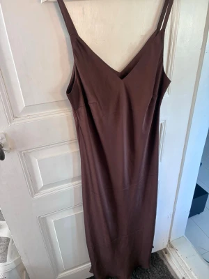 Brun långklänning med smala axelband - Säljer en stilren brun långklänning med smala axelband och v-ringning från Lindex. Klänningen har en rak passform och är perfekt för dig som gillar minimalistisk stil. Materialet känns mjukt och följsamt mot kroppen.