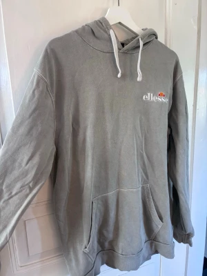 Grå hoodie från Ellesse - Snygg grå hoodie från Ellesse med vit dragsko i huvan och klassisk logga broderad på bröstet. Hoodien har en stor magficka och ribbade muddar vid ärmslut och nederkant. Perfekt för en chill och avslappnad stil.