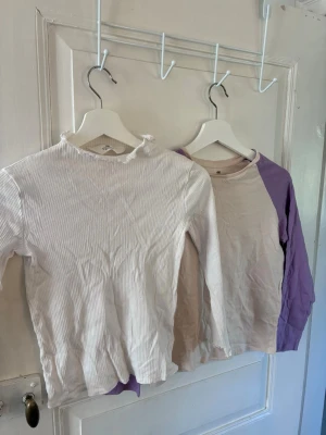 Två långärmade toppar för flickor från H&M - Säljer två långärmade toppar för flickor från H&M. Ena toppen är vit och ribbad med liten volangkant vid halsen, den andra är beige med lila raglanärmar. Båda är i mjuk bomull och har en normal passform. Perfekta för en chill och avslappnad stil.