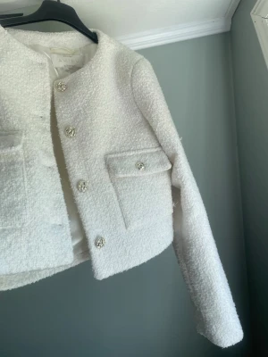 Vit bouclé kavaj från Amisu - Stilren vit bouclé kavaj från Amisu med dekorativa guldknappar och två fickor framtill. Kortare modell med rundad halsringning och fodrad insida. Perfekt för en chic och trendig look.
