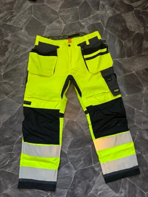 Snickers varselbyxa gul/svart - Helt ny.  Säljs pga att jag köpte fel storlek  Varselbyxa från Snickers i high vis gul och svart med grå detaljer. Byxan har flera praktiska fickor, förstärkta knän och reflexband för extra synlighet. Perfekt för dig som jobbar inom bygg eller industri och vill ha både funktion och stil.