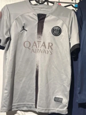 Grå PSG matchtröja Jordan - Snygg grå Paris Saint-Germain matchtröja i samarbete med Jordan. Tröjan har en svart vertikal rand i mitten, PSG-logga på bröstet och Jordan-logga på andra sidan. Qatar Airways som sponsortryck på framsidan. Tillverkad i lätt och ventilerande material. Neymar 10 på ryggen