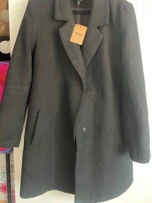 Svart klassisk kappa från See by - Stilren svart kappa med enkel knäppning och klassisk krage från See by. Kappan har raka ärmar, två sidofickor och en rak passform. Perfekt för dig som gillar minimalistisk och tidlös stil. Materialet ser ut att vara ullblandning.