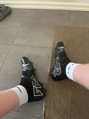 Svarta Puma fotbollsskor med socka - Säljer ett par svarta fotbollsskor från Puma med vit logga och FG-dobbar. Skorna har en strumpliknande krage för extra stöd runt ankeln och en modern design med vita detaljer på sidan. Perfekta för dig som vill ha stilrena och bekväma skor på planen.