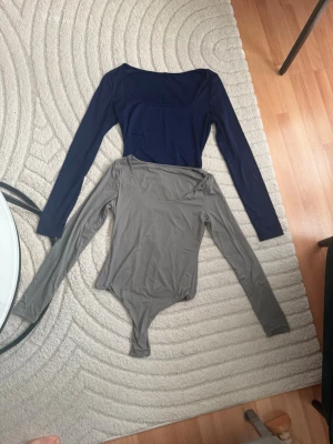 Två långärmade bodys i blått och grått - Säljer två stilrena långärmade bodys med rundad urringning, en i marinblått och en i ljusgrått. Båda är i mjukt, stretchigt material som sitter snyggt på kroppen. Perfekta att matcha med jeans eller kjol för en clean look.