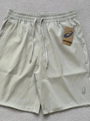 Asics träningsshorts med snörning - Snygga träningsshorts från Asics i svart och ljusbeige. De har elastisk midja med snörning och Asics-logga på benet. Perfekta för sport eller gym och tillverkade i lätt syntetmaterial som andas. Olika storlekar finns!