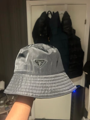 Svart bucket hat från Prada - Snygg svart bucket hat från Prada i glansigt nylonmaterial. Klassisk form med bred skärm och ikoniska metalltriangeln med logga framtill. Perfekt accessoar för en trendig streetwear-look.