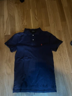 Marinblå pikétröja Polo Ralph Lauren - Klassisk marinblå pikétröja från Polo Ralph Lauren i slim fit-modell. Tröjan har korta ärmar, krage och knappar framtill samt den ikoniska röda loggan broderad på bröstet. Perfekt till jeans eller shorts för en clean look.