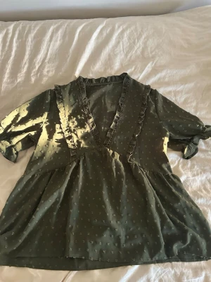 Olivgrön blus med volangdetaljer - Supersöt olivgrön blus med små diskreta prickar och volang längs den djupa v-ringningen. Blusen har trekvartsärm med lätt puff och resår vid ärmslutet. Materialet är tunt och flowy, perfekt för en trendig look. Passar vem som helst det bror på vad man satsar på för look