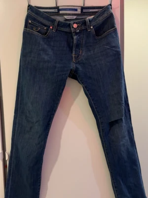Jacob cohen jeans - Säljer ett par mörkblå jeans från Nudie Jeans med klassisk femficksmodell och raka ben. Jeansen har en patchad detalj på höger ben och råa, avklippta benslut för en trendig look. Tillverkade i denim med kontrastsömmar och knappgylf.