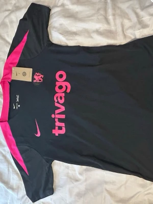 Chelsea Nike Dri-Fit svart tränings-tshirt - Snygg svart tränings-tshirt från chelsea med rosa detaljer och tryck. Tröjan har Dri-Fit-material som andas, rosa logga och texten 'trivago' på bröstet. Perfekt för fotboll eller gym. Modern passform och sportig look.
