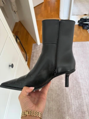 Svarta boots med smal klack - Snygga svarta stövletter i slätt skinnimitation med spetsig tå och smal klack. Dragkedja på sidan för enkel på- och avtagning. Perfekta till jeans eller kjol. Dom är endast använda en gång och behöver bara torkas av med en blöt trasa vilket jag kommer göra innna jag skickar dom. Dom är i storlek 35 men skulle även passa 36