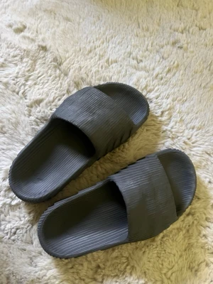 Adidas Adilette 22 slides - Säljer ett par mörkgrå adidas tofflor i ribbad design med bred rem över foten. Perfekta för att glida runt hemma eller på stranden.