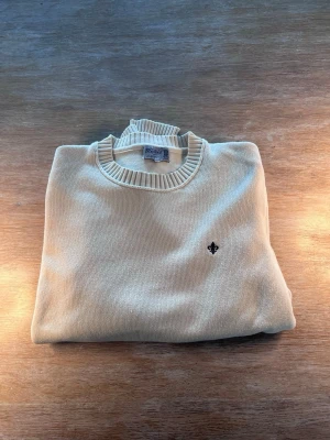 Beige stickad tröja Morris - Klassisk beige stickad tröja från Morris med rund halsringning och ribbade muddar vid ärmslut och nederkant. Tröjan har en liten svart broderad logga på bröstet och är tillverkad i mjukt material som passar perfekt till lager-på-lager.
