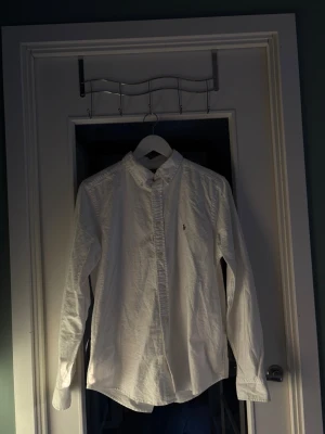 Vit skjorta från Ralph Lauren - Klassisk vit skjorta från Ralph Lauren med button-down krage och broderad logga på bröstet. Skjortan har lång ärm och knäppning framtill. Perfekt för en clean och stilren look.