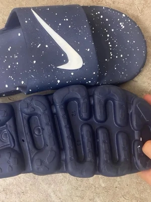 Blå Nike tofflor med vit stänkmönster - Säljer ett par blåa Nike tofflor med vit stänkmönster och stor vit Swoosh-logga på ovandelen. Tillverkade i syntetmaterial med mönstrad sula för extra grepp. Perfekta för chill dagar hemma eller efter träningen.
