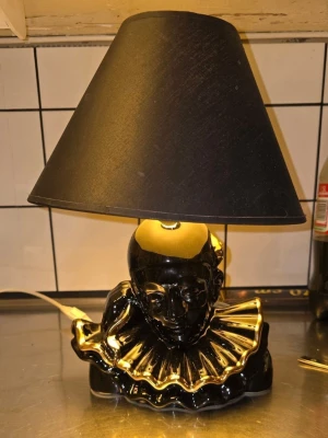 Svart bordslampa med clownmotiv (80tal) - Unik bordslampa i svart keramik med motiv av en clownbyst och detaljer i guld. Lampan har en svart lampskärm och en dekorativ krage med guldkant. Perfekt som inredningsdetalj för dig som gillar retro eller udda design.köpt på 1980 talet