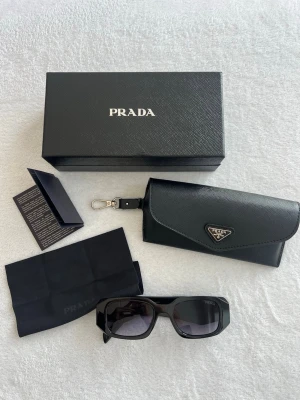 Svarta solglasögon från Prada - Snygga svarta solglasögon från Prada med breda skalmar och rektangulär form. Glasögonen har tydlig Prada-logga på sidan och kommer med originalfodral, putsduk och box. Perfekt accessoar för en trendig look.