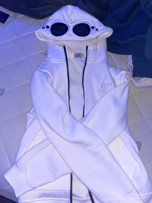 Vit hoodie med goggles från C.P. Company - Säljer en vit hoodie från C.P. Company med ikoniska inbyggda goggles i huvan och dragkedja framtill. Märkets logga är broderad på bröstet. Hoodien har en clean och modern look, perfekt för dig som vill sticka ut. Materialet känns mjukt och bekvämt.