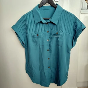 Turkos blus med kort ärm och fickor - Stilren turkos skjorta med korta ärmar och uppvik, knäppning framtill och två bröstfickor med dekorativa sömmar. Skjortan har en klassisk krage och ribbade detaljer upptill. Perfekt att matcha med jeans för en avslappnad look. Storlek L. Från armhålan till armhålan 54cm. Oanvänd. 
