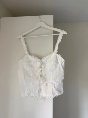Vit snörad linnetopp från Gina Tricot - Supersöt vit linnetopp från Gina Tricot med snörning framtill och spetsdetaljer längs kanterna. Toppen har breda axelband och en croppad passform som ger en romantisk vibe. Perfekt att styla till jeans eller kjol för en trendig look.