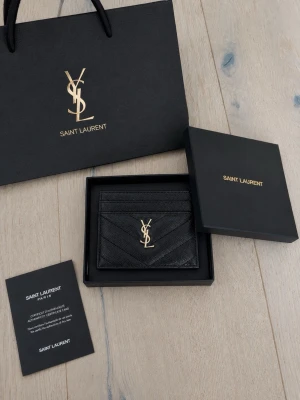 Svart korthållare från Saint Laurent - Snygg svart korthållare från Saint Laurent i läder med guldfärgad YSL-logga framtill. Klassisk chevronmönstrad design och flera fack för kort. Perfekt accessoar för dig som gillar stilrena detaljer och lyxig känsla.
