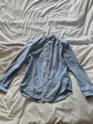 Blå skjorta från Ralph Lauren - Klassisk ljusblå skjorta från Ralph Lauren med broderad logga på bröstet. Skjortan har knappar framtill, button-down krage och långa ärmar. Tillverkad i mjuk bomull som känns skön mot huden. Perfekt för en clean och stilren look.