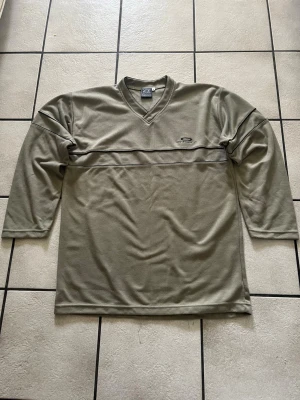 Olivgrön Oakley tröja med V-ringning storlek L, bomull  - Snygg olivgrön långärmad tröja från Oakley med V-ringad hals och diskret broderad logga på bröstet. Tröjan har svarta detaljer i form av tunna linjer över bröstet och är tillverkad i ett mjukt, ribbat material.