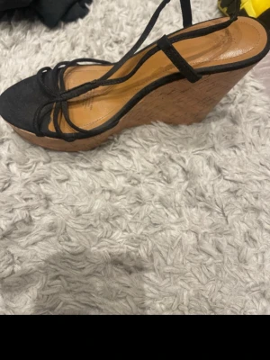 Svarta kilklackade sandaler - Säljer ett par svarta sandaler med höga kilklackar i kork. Skorna har tunna remmar över foten och öppen tå. Perfekta för dig som vill ha både höjd och stil. Innersulan är i brunt konstläder och yttersulan är i korkmaterial.