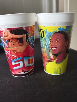 Färgglada plastmuggar med fotbollstema - Två plastmuggar med färgstarka illustrationer av fotbollsspelare och grafiska element. Muggarna har vita kanter och motiv i rött, blått, gult och grönt. Perfekta till kalas eller som samlarprylar för fotbollsälskare.