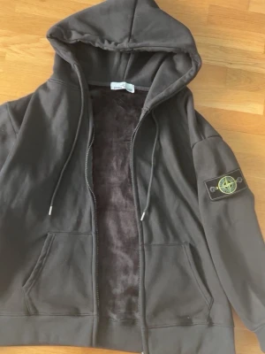 Svart hoodie från Stone Island - Svart hoodie med dragkedja från Stone Island. Klassisk patch med logga på vänster ärm, huva med dragsko och två stora fickor framtill. Mjuk insida och ribbade muddar. Perfekt för en streetwear-look.