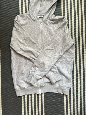 Grå zip hoodie från Kappahl - En enkel och stilren grå hoodie från Kappahl med dragkedja framtill och två fickor. Hoodien har ribbade muddar vid ärmslut och nederkant samt en klassisk huva. Perfekt för en chill och avslappnad look. Storlek 158/164.