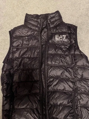 Svart quiltad dunväst från EA7 Armani - Snygg svart dunväst från EA7 Emporio Armani med quiltad design och hög krage. Västen har dragkedja framtill och en stilren logga på bröstet. Perfekt för lager på lager och ger en sportig vibe till din outfit.
